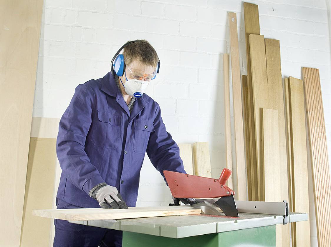 Muž v ochranném oblečení pracuje s kotoučovou pilou v dílně. V pozadí jsou naskládané dřevěné desky.