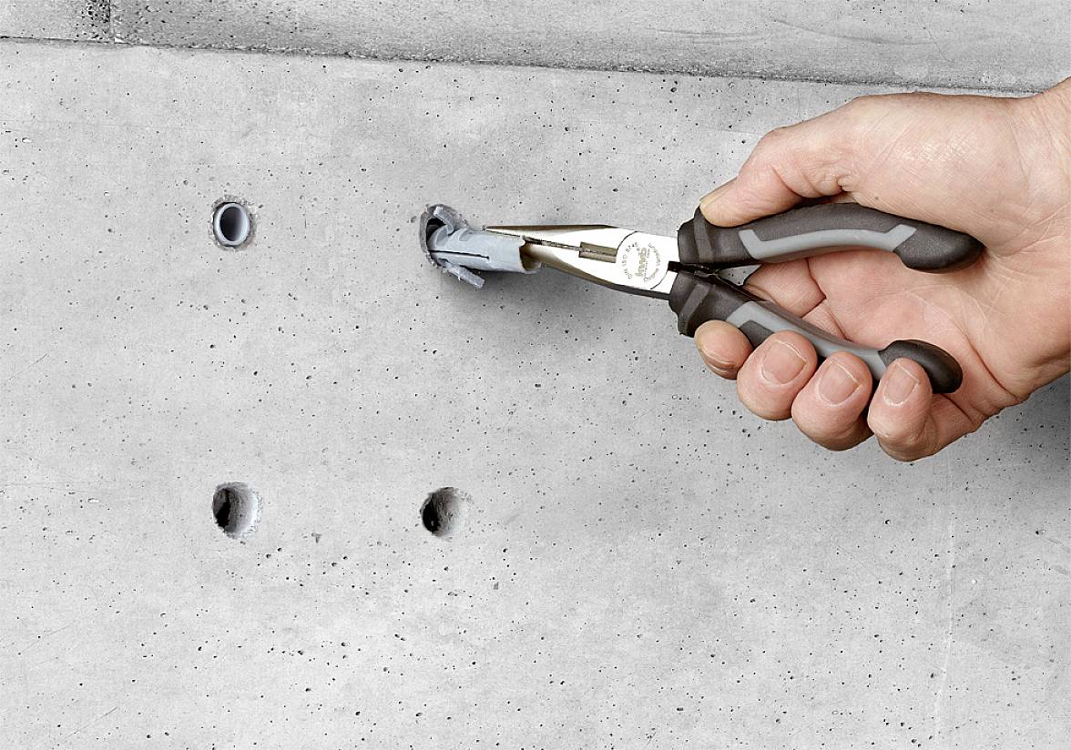 Jedna ruka vytahuje pomocí kleští hmoždinku z betonové stěny. Vedle jsou viditelné další otvory.