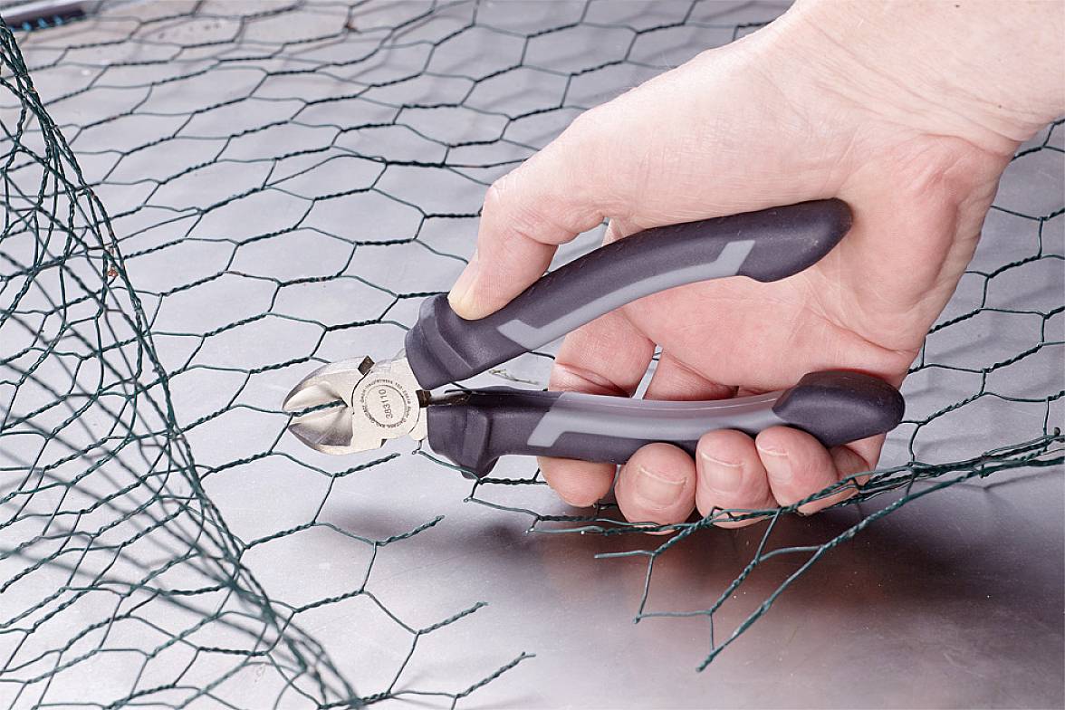 Jedna ruka řeže drátěným štípacím kleštěmi plot z pletiva. Drát je stříhán na kovovém povrchu.