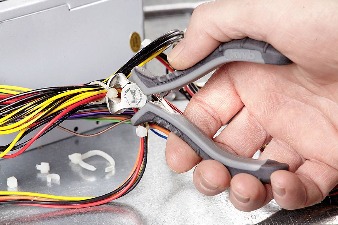 Jedna ruka drží boční štípací kleště a stříhá dráty. V elektronickém krytu je několik barevných kabelů.