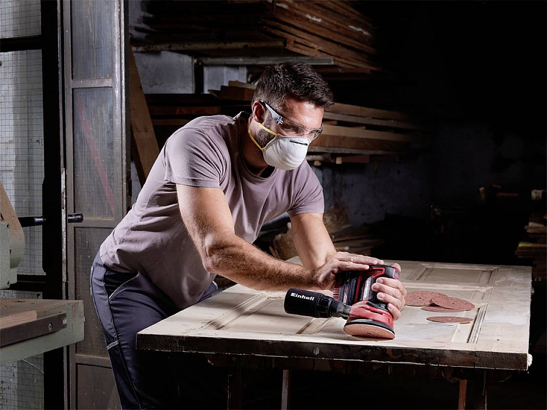 Muž nosí respirátor a ochranné brýle, zatímco broušením dřevěné desky elektrickou bruskou na pracovním stole.