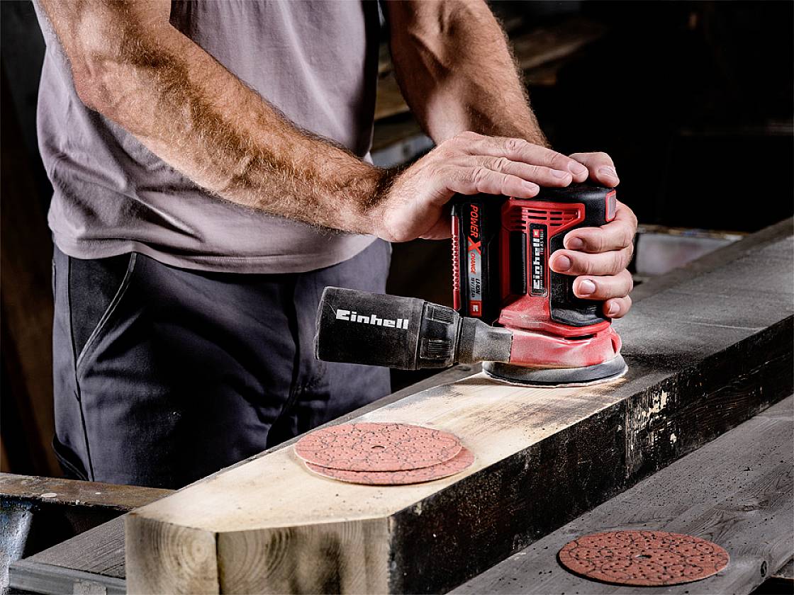 Osoba brousí pomocí brusky Einhell na dřevěném stole. Jsou vidět také brusný papír a kus dřeva.