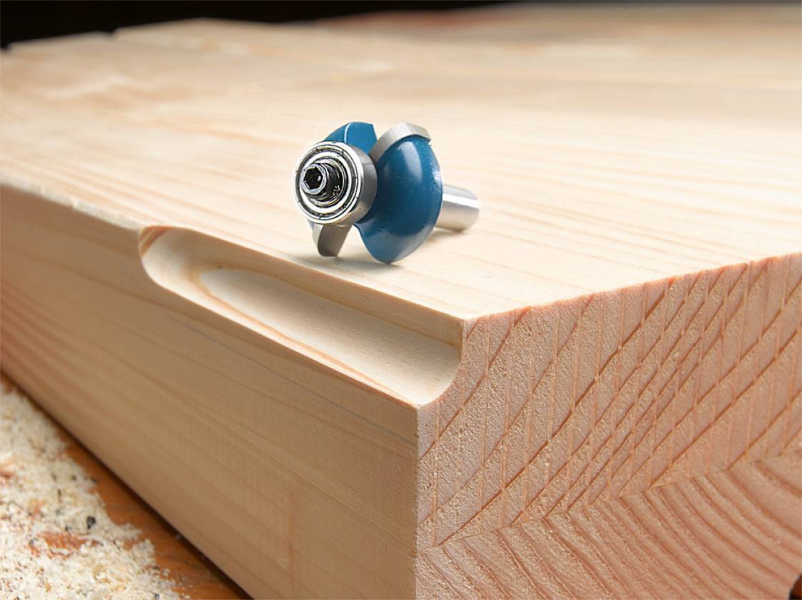 Dřevěný kus s frézovací kapsou, na němž leží fréza, obklopený dřevěnými pilinami.