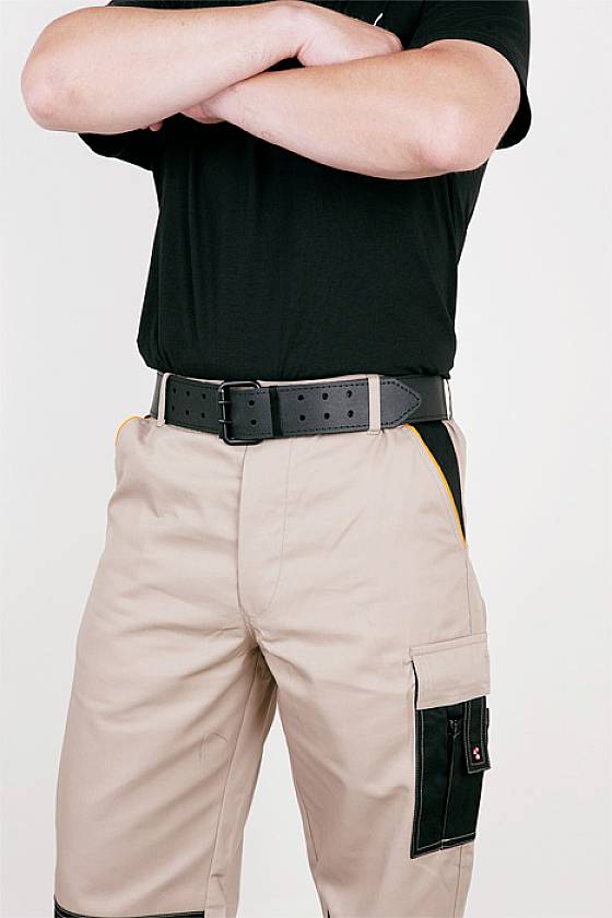 Osoba v béžových pracovních kalhotách s černým páskem. Paže má překřížené, košile je černá. Nerozpoznatelné obličejové rysy.