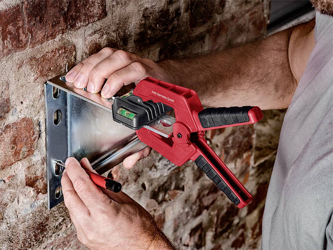 Osoba upevňuje kovovou desku na cihlovou stěnu pomocí červené svěrky, používá vodováhu a tužku k vyznačení polohy.