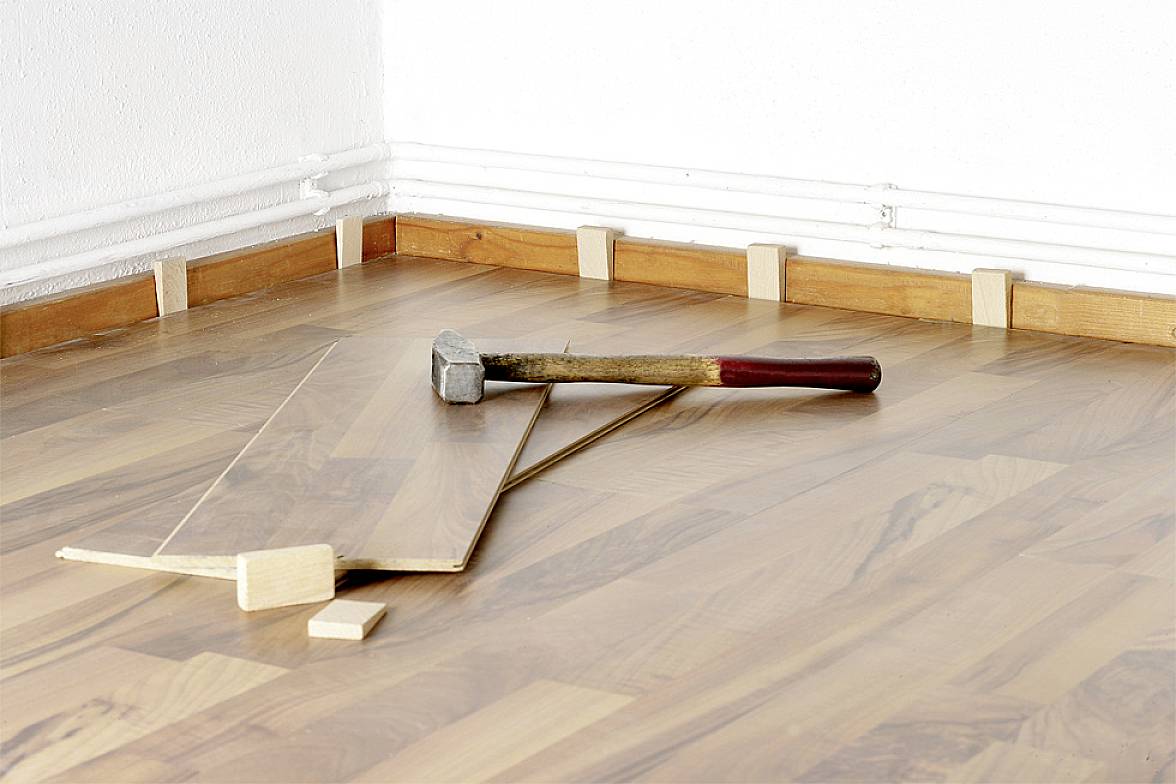 Na parketové podlaze leží kladivo s volnou dřevěnou podlahovou deskou a distančními klíny u stěny.