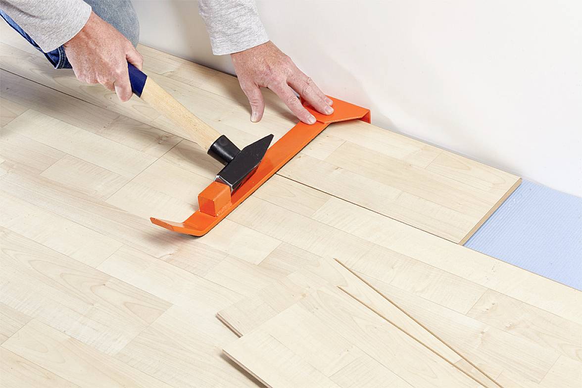 Osoba instaluje laminátovou podlahu pomocí kladiva a oranžového instalačního nástroje pro úpravu panelů.