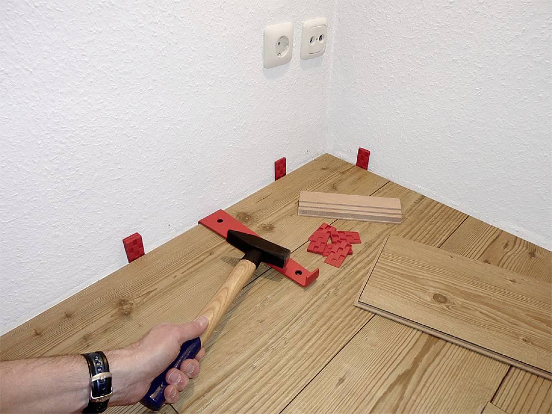 Osoba pokládá laminátovou podlahu. Kladivo se používá k upevnění desky na okraji. Distanční klínky jsou umístěny u stěny.