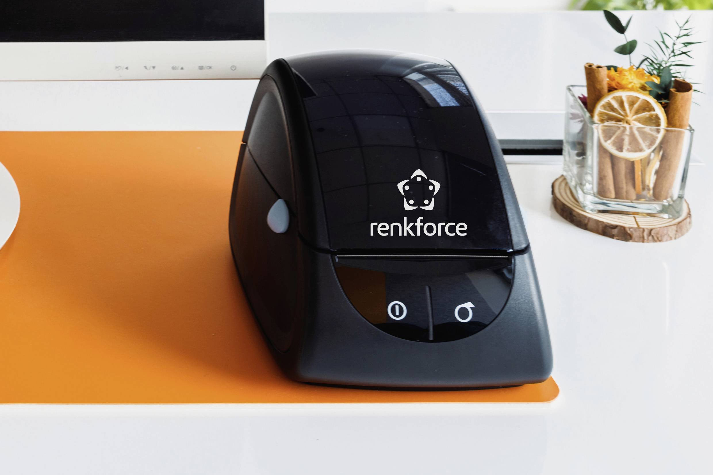 Černá tiskárna štítků značky Renkforce stojí na oranžovém podkladu, vedle ní dekorace ze skořice a citronu.