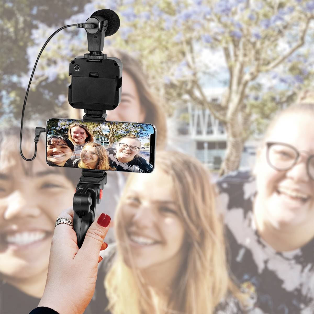 Jedna ruka drží smartphone s mikrofonem, na kterém je vidět selfie čtyř usmívajících se osob. Pozadí je rozmazané.