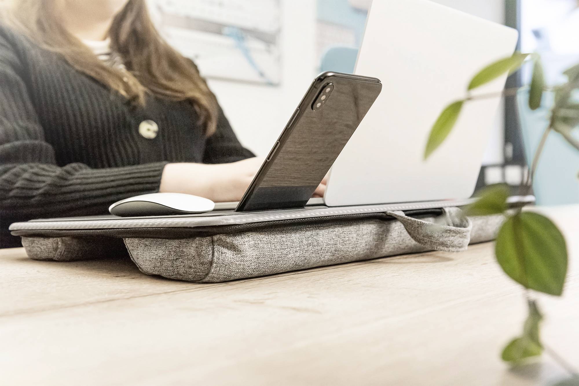 Žena pracuje na laptoptpu s mobilním telefonem a myší na šedém podložce pod notebook. V popředí je viditelná rostlina.