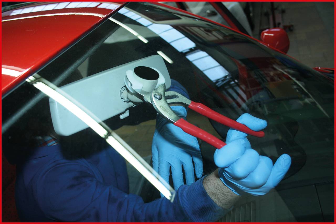 Mechanik v modrých rukavicích používá kleště k upevnění zpětného zrcátka na čelním skle červeného automobilu.