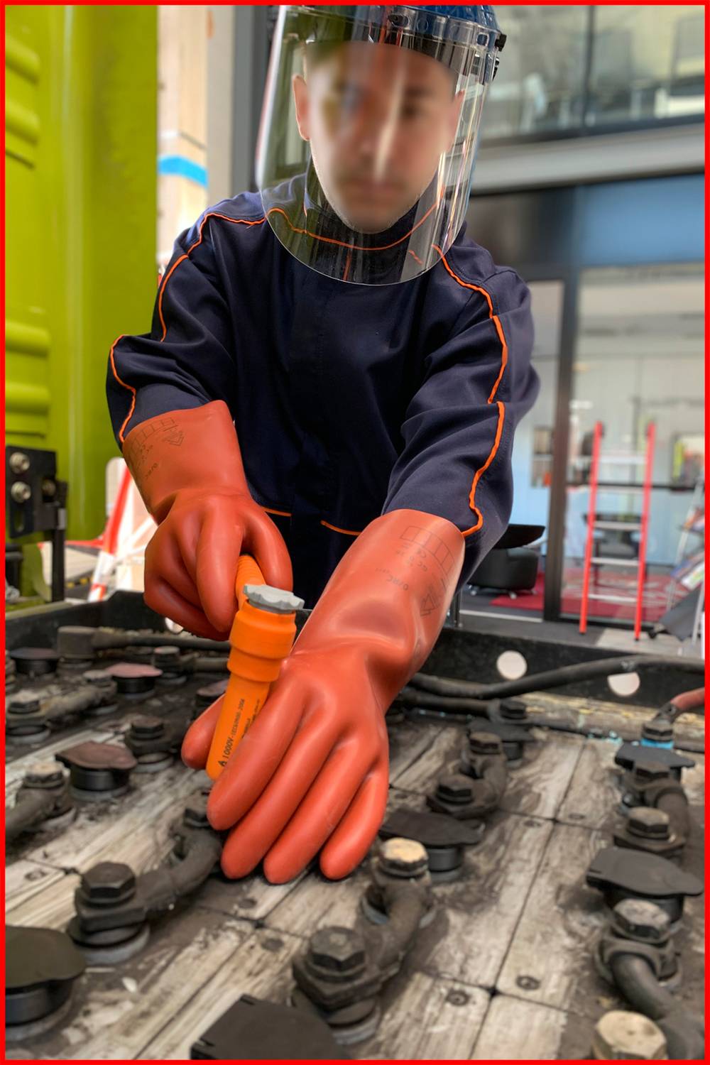 Osoba v ochranném oblečení a rukavicích pracuje s nástroji na velkých bateriích v průmyslovém prostředí.