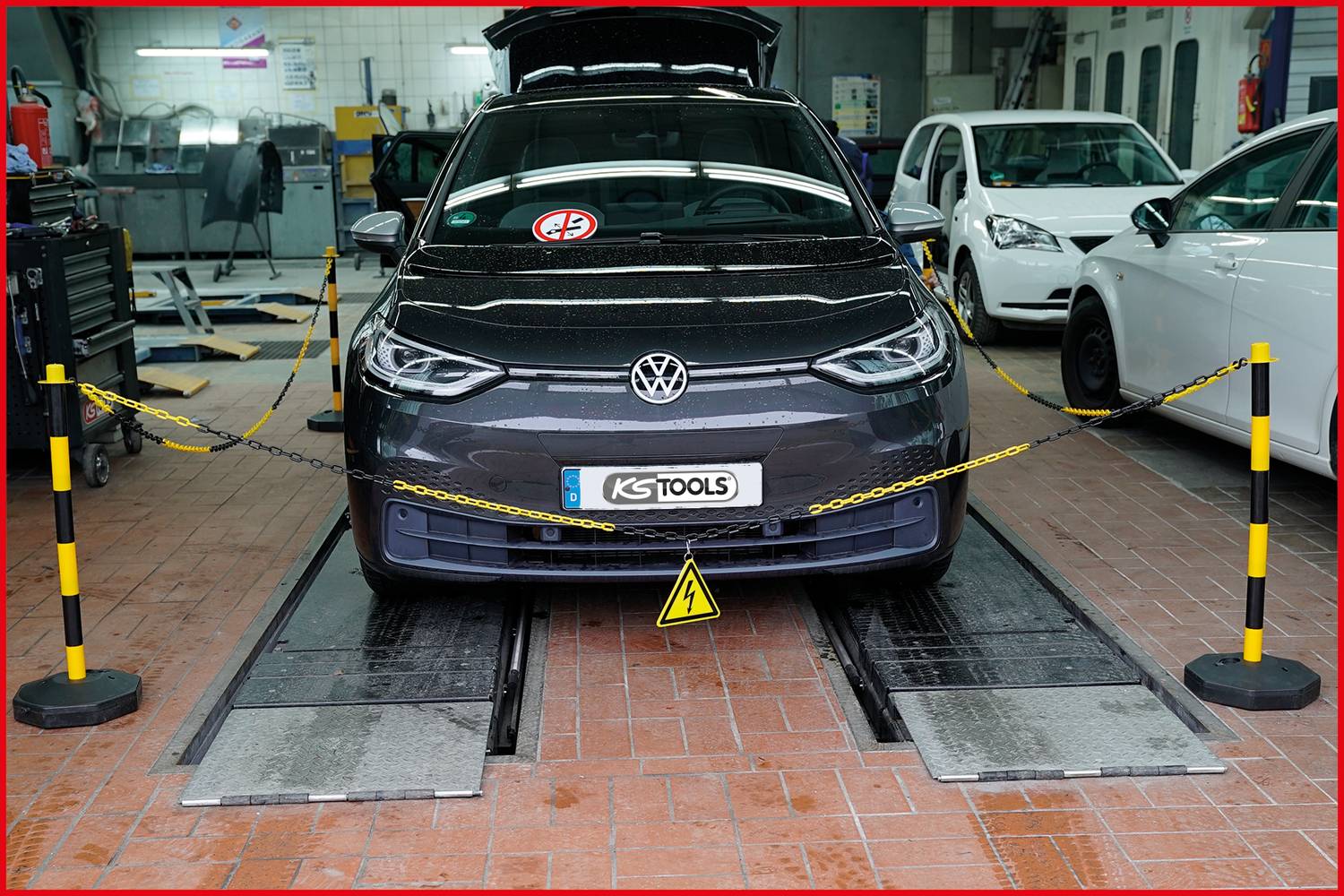 Šedé auto stojí v dílně na zvedáku. Je obklopeno bezpečnostními řetězy a dalšími vozidly v pozadí.