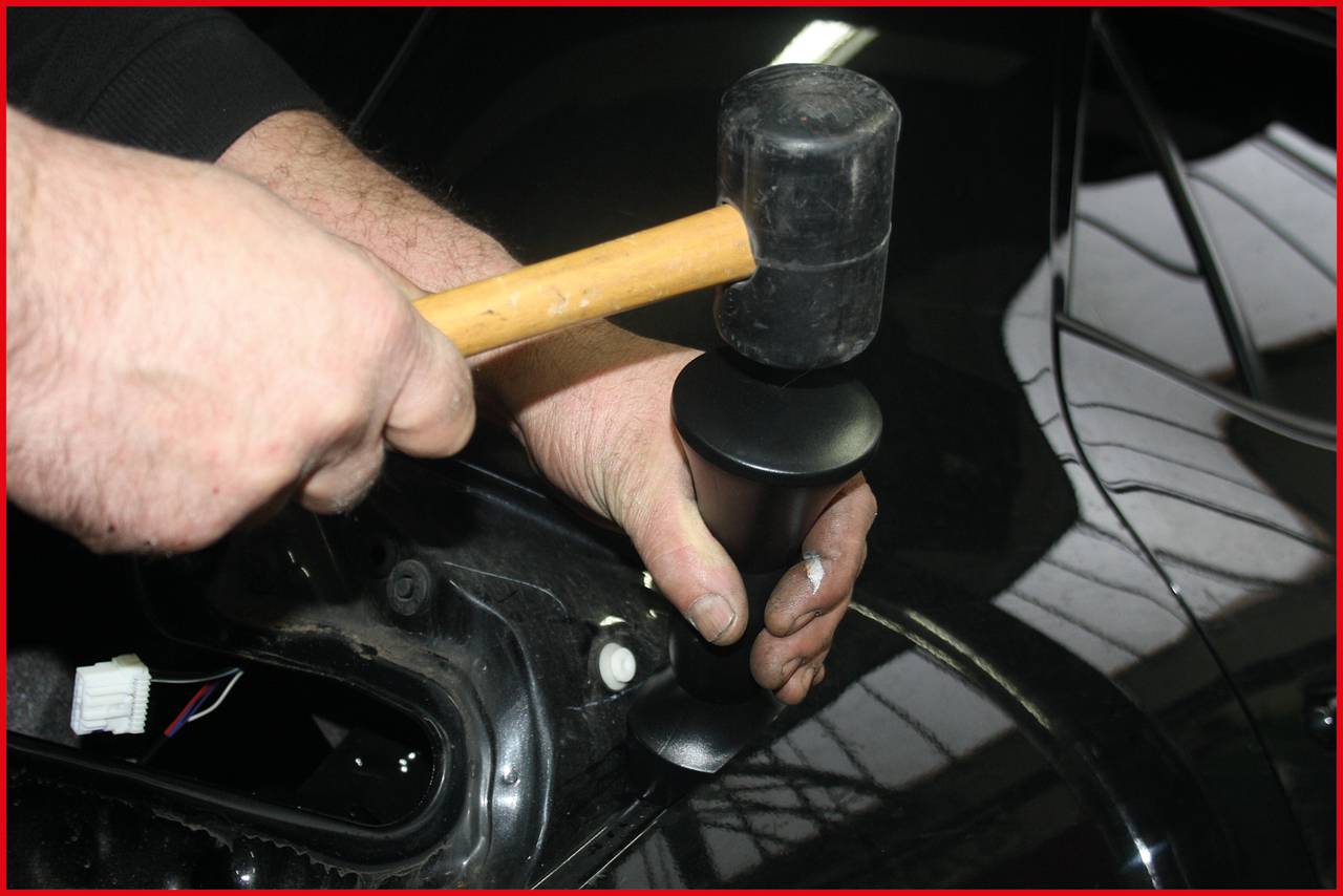Osoba opravuje plech automobilu gumovou palicí. Prostředí je automechanická dílna.