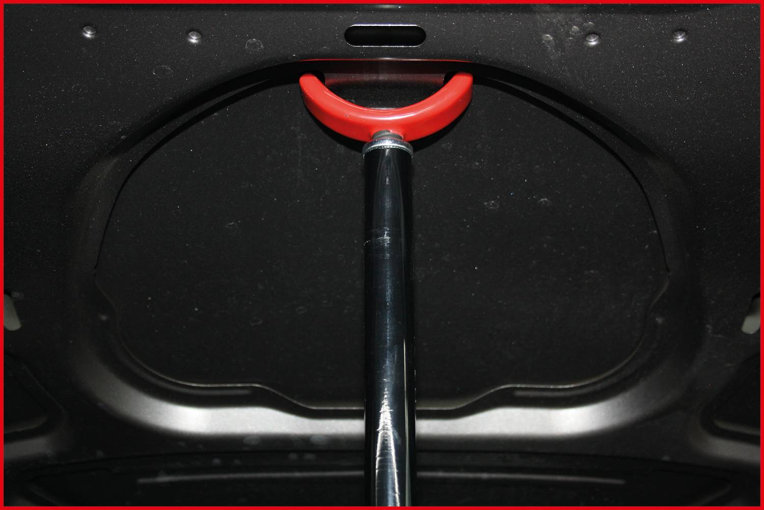 Kapota automobilu s podpěrnou tyčí uprostřed, opatřená červeným madlem pro zvedání nebo stabilizaci otevřené kapoty.