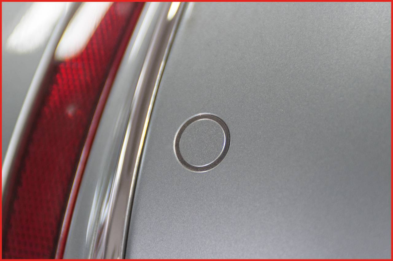 Detailní pohled na šedý nárazník automobilu s kulatým, zapuštěným senzorem umístěným vlevo vedle červené zadní světlo.