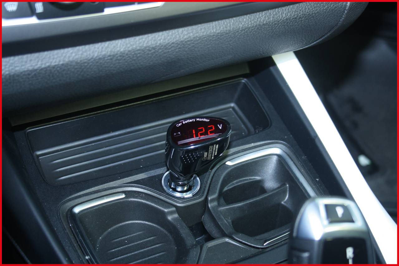 Měřič napětí v zapalovači automobilu ukazuje '12,2 V'. Vedle jsou umístěny dva prázdné držáky nápojů.