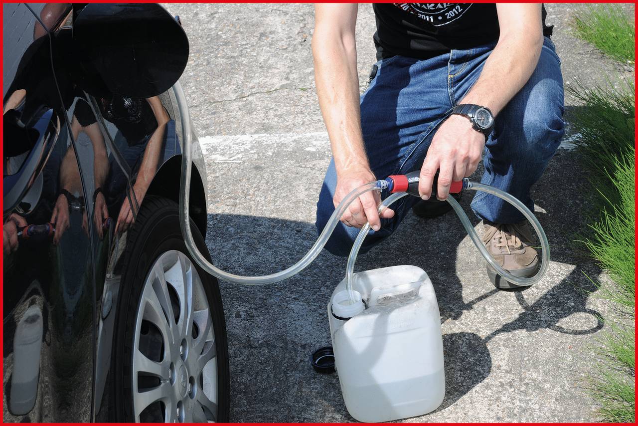 Osoba nalévá hadičkou benzín z bílého kanystru do automobilu. Venkovní scéna s cestou a trávou v pozadí.