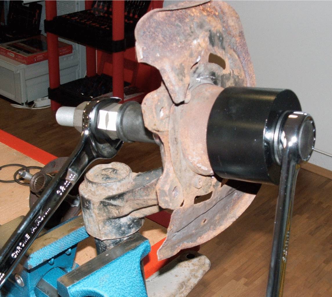 Mechanismus nářadí pracující na rezavém kovovém kotouči, upnutém ve svěráku, obklopený vybavením dílny.