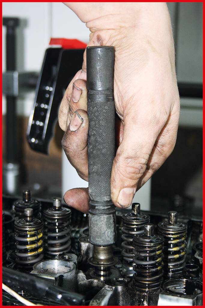 Jedna ruka drží nástroj nad otevřeným motorem s viditelnými pružinami a mechanikou.