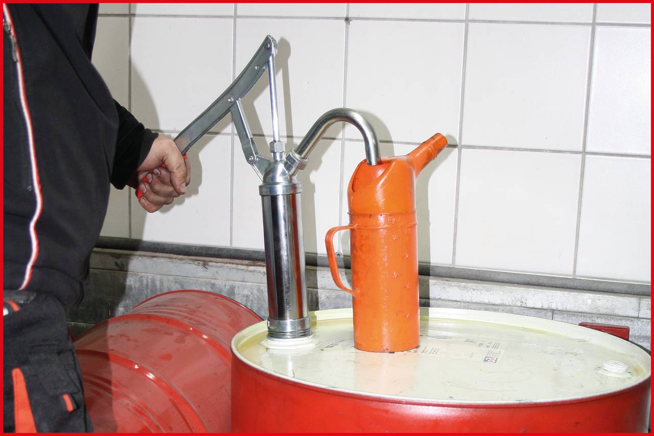 Člověk čerpá kapalinu z velké červené barely pomocí ruční pumpy. Vedle pumpy stojí oranžová olejová nádoba.
