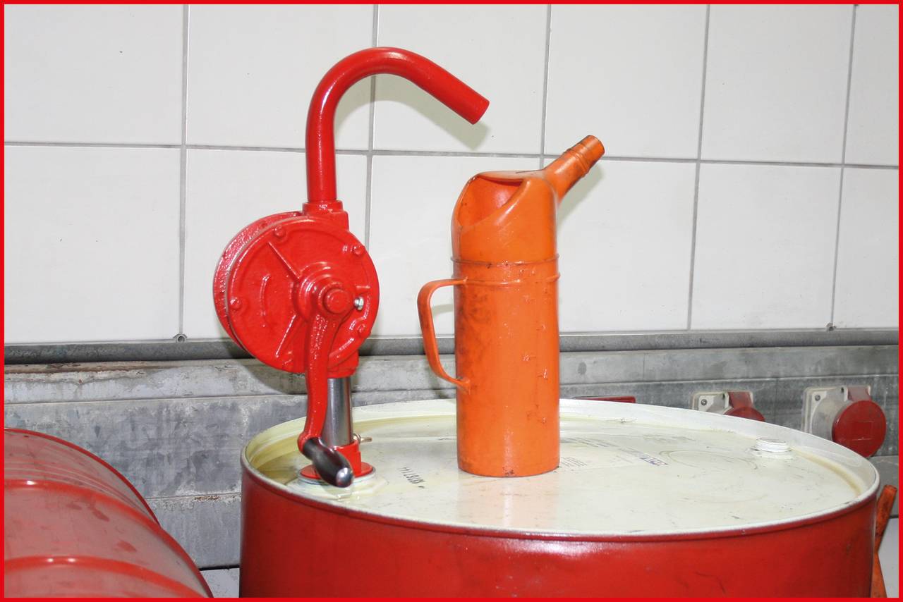 Červená ruční pumpa na sudu, vedle oranžový kanystr. Zařízení se nachází v místnosti s obloženou stěnou v pozadí.
