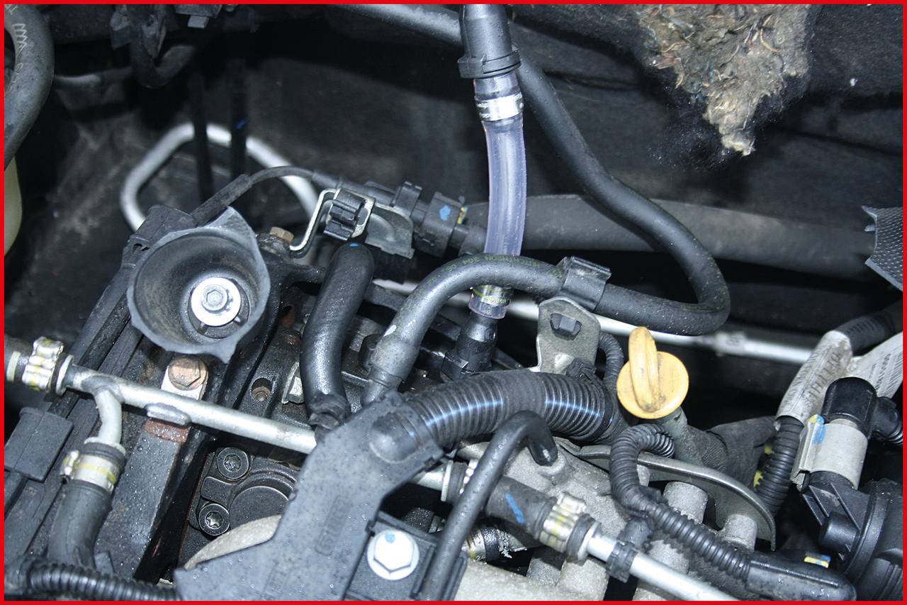 Motorový prostor s hadicemi a kabely. Žlutá měrka oleje je viditelná obklopená různými komponenty vozidla.