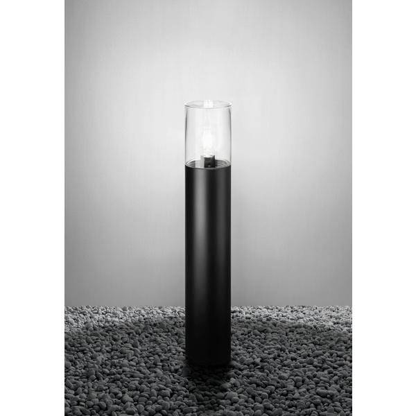 Volně stojící černá kovová lampa s průhledným skleněným stínidlem, stojící na štěrkovém povrchu, rozsvícená před šedým pozadím.