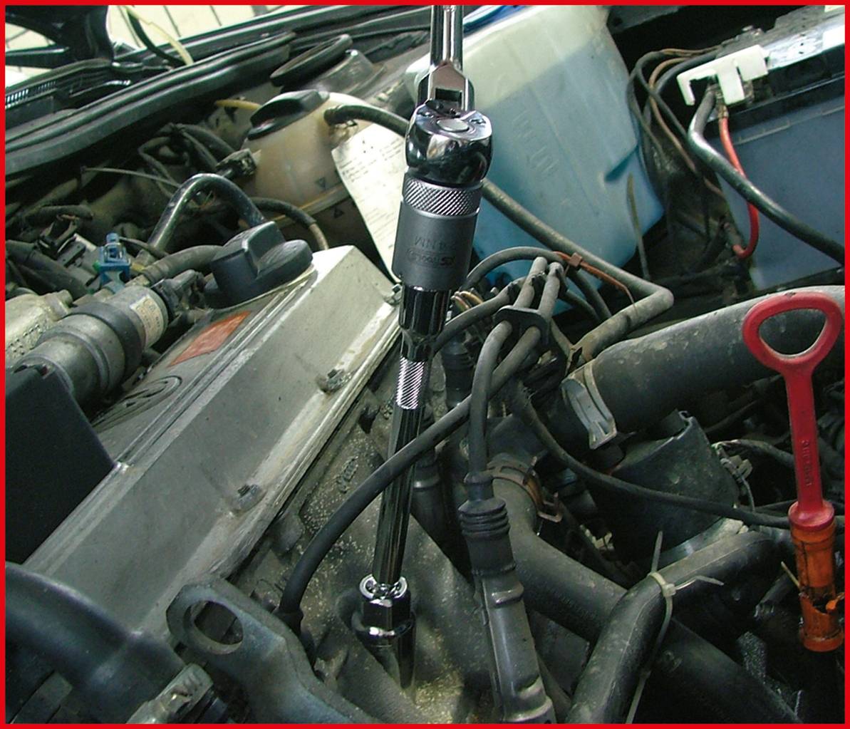 Momentový klíč se používá na motoru automobilu. Kolem klíče jsou viditelné různé motorové komponenty a nástroje.