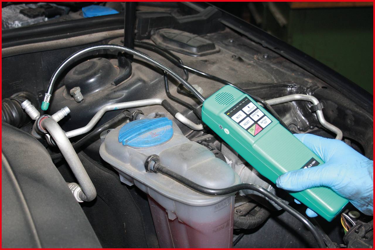 Osoba používá elektronické zařízení k vyhledávání netěsností v motorovém prostoru vozidla, obklopena hadicemi a kryty.