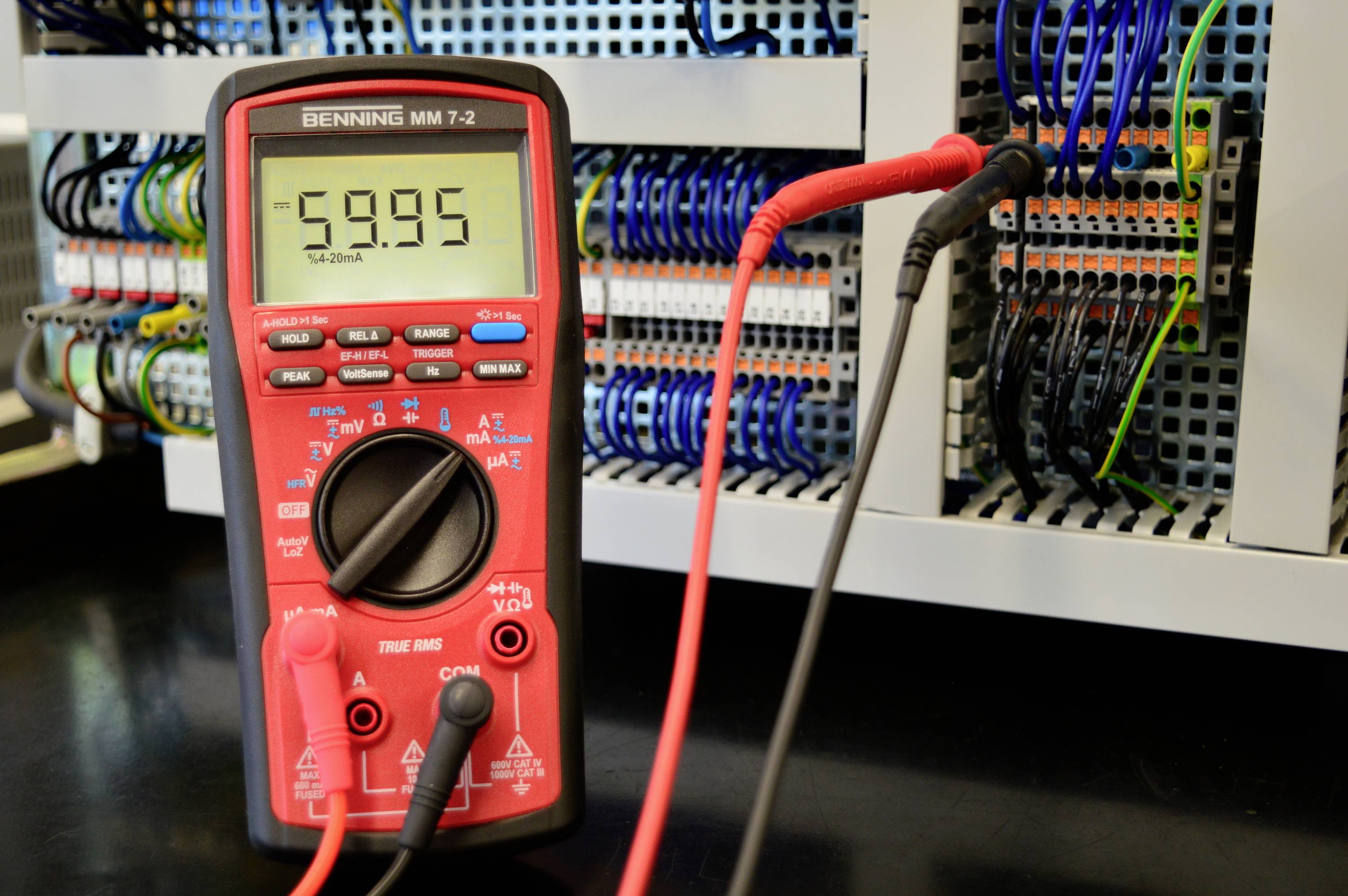 Červený multimetr měří -59,95 na rozvodné desce s mnoha vodiči, symbolizuje měření v elektronice a elektrotechnice.