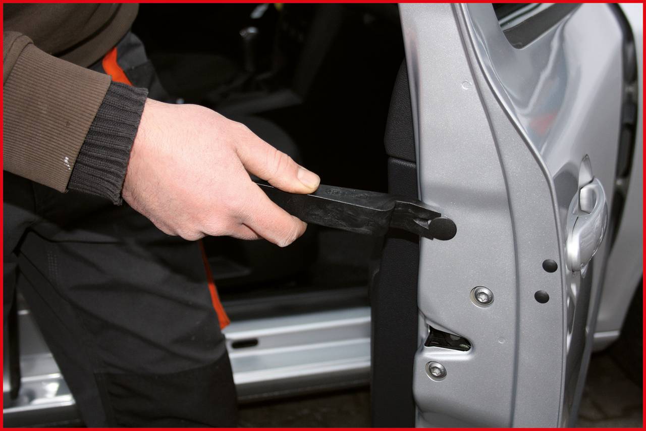 Osoba používá páčicí nástroj k otevření automobilu bez poškození. Autovýklopné dveře jsou lehce pootevřené a osoba má na sobě pracovní oděv.