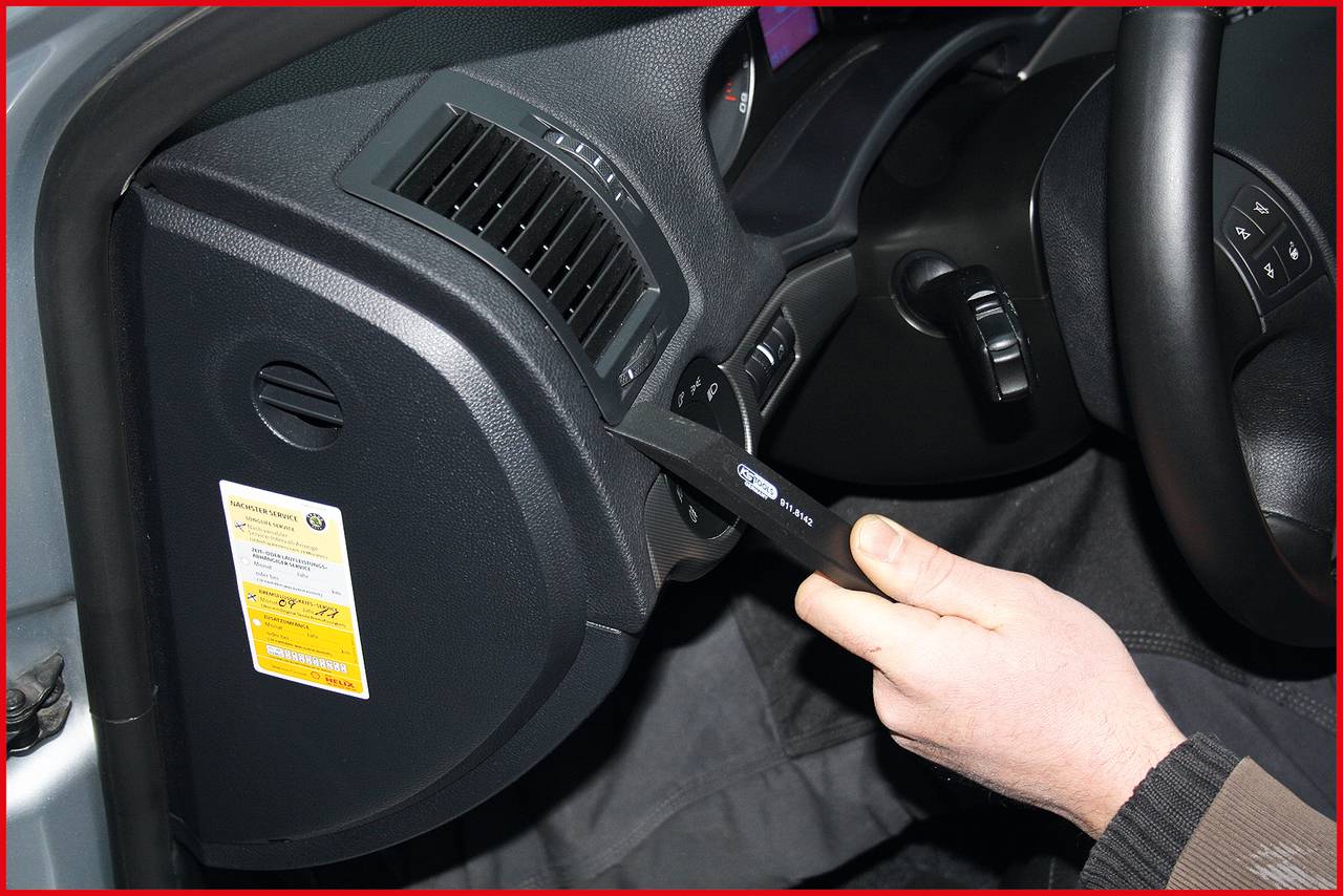 Ruka drží nástroj, který otevírá malou přihrádku na straně automobilu. Ovládací tlačítka a větrací štěrbiny jsou viditelné.