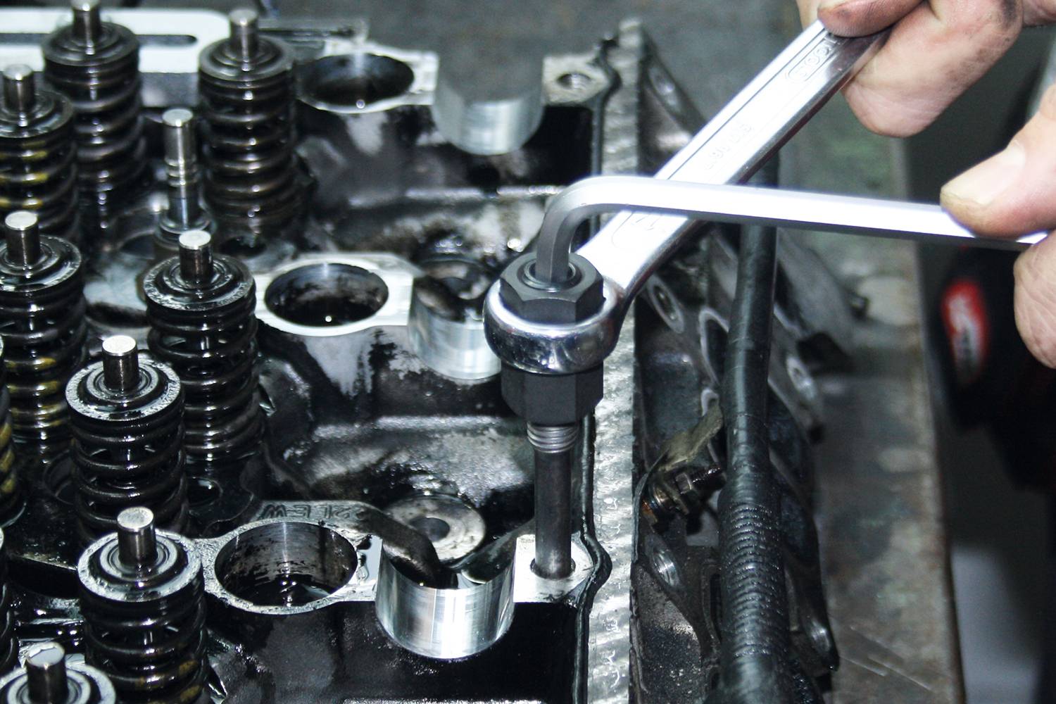 Mechanik utahuje šroub na bloku motoru pomocí klíče. Několik ventilových pružin je viditelných.