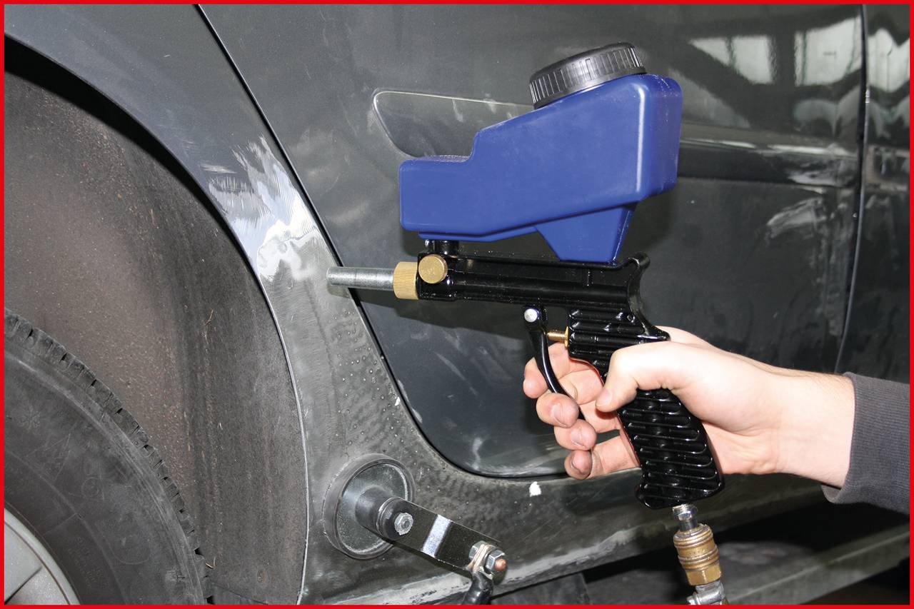 Osoba drží stříkací pistoli a pracuje na opravě automobilu. Na straně vozidla jsou patrné stopy po broušení.