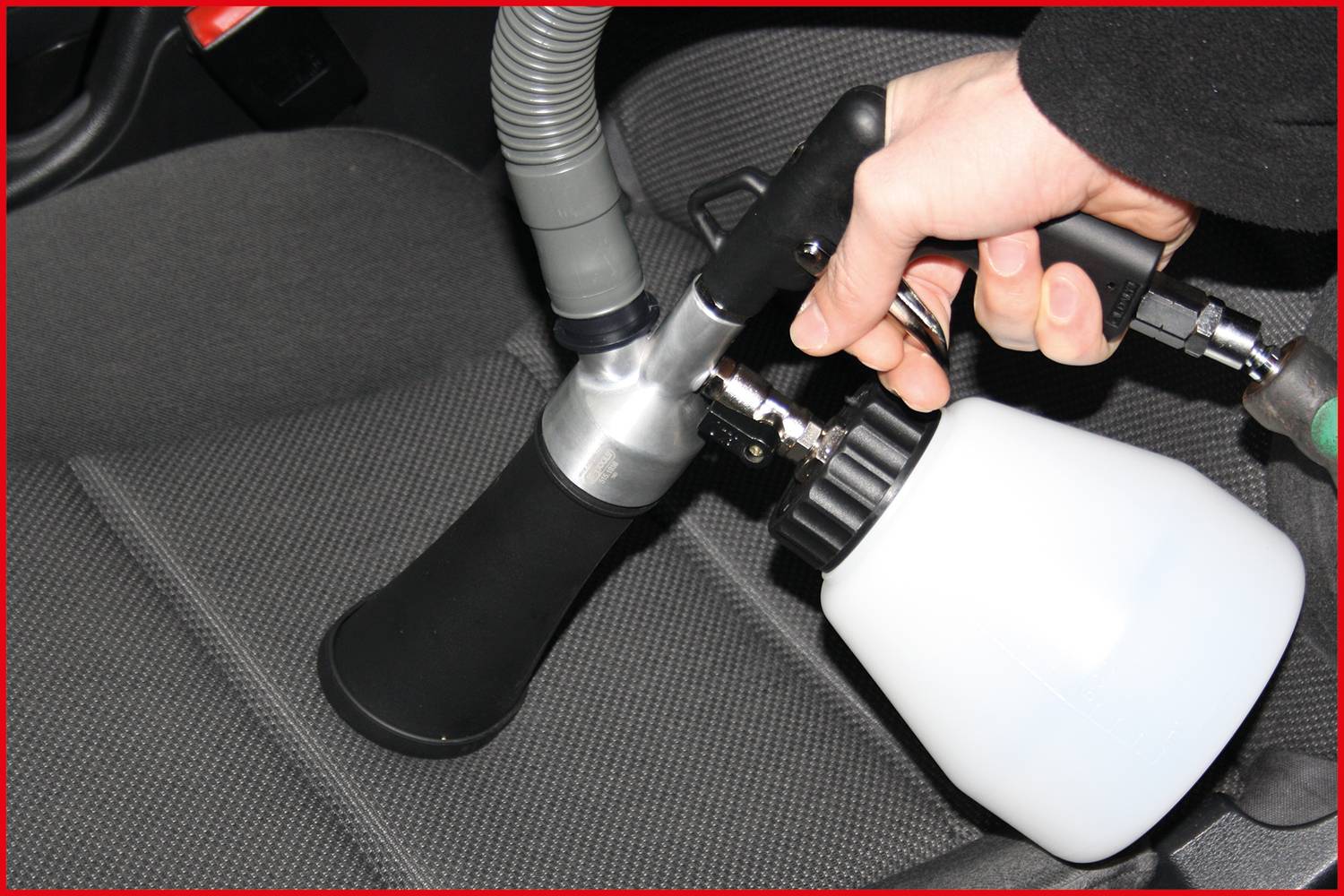 Ruka drží čisticí nástroj na sedadle automobilu, připravena k použití. Bílá láhev je součástí zařízení. Sedadlo je potaženo šedou látkou.