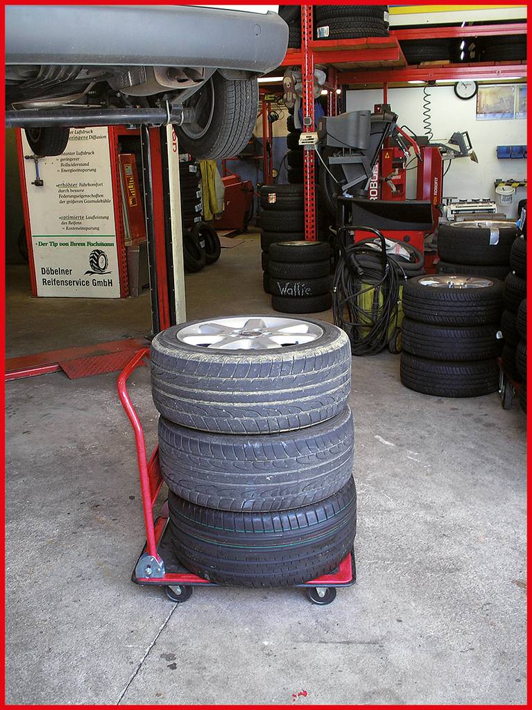 Pneumatiky na voze v autoservisu. Zvedák s nadzvednutým autem v pozadí, další pneumatiky a vybavení jsou viditelné.