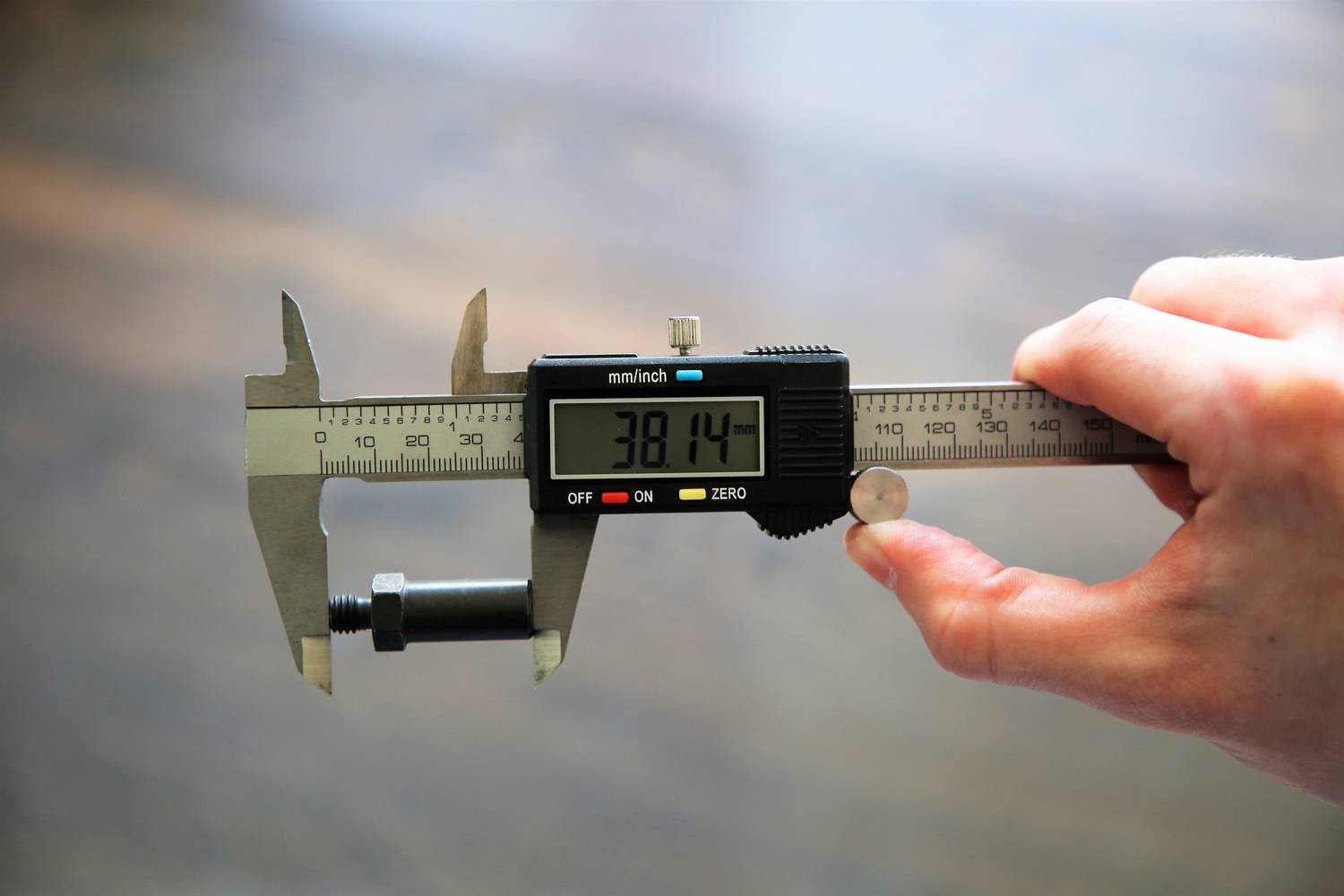 Jedna ruka drží digitální posuvné měřidlo, které na displeji zobrazuje délku malého šroubu jako '38,14 mm'.