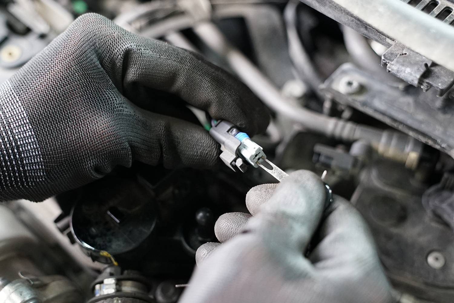 Osoba v černých rukavicích kontroluje malou automobilovou součástku v motorovém prostoru. Probíhá mechanická údržba.