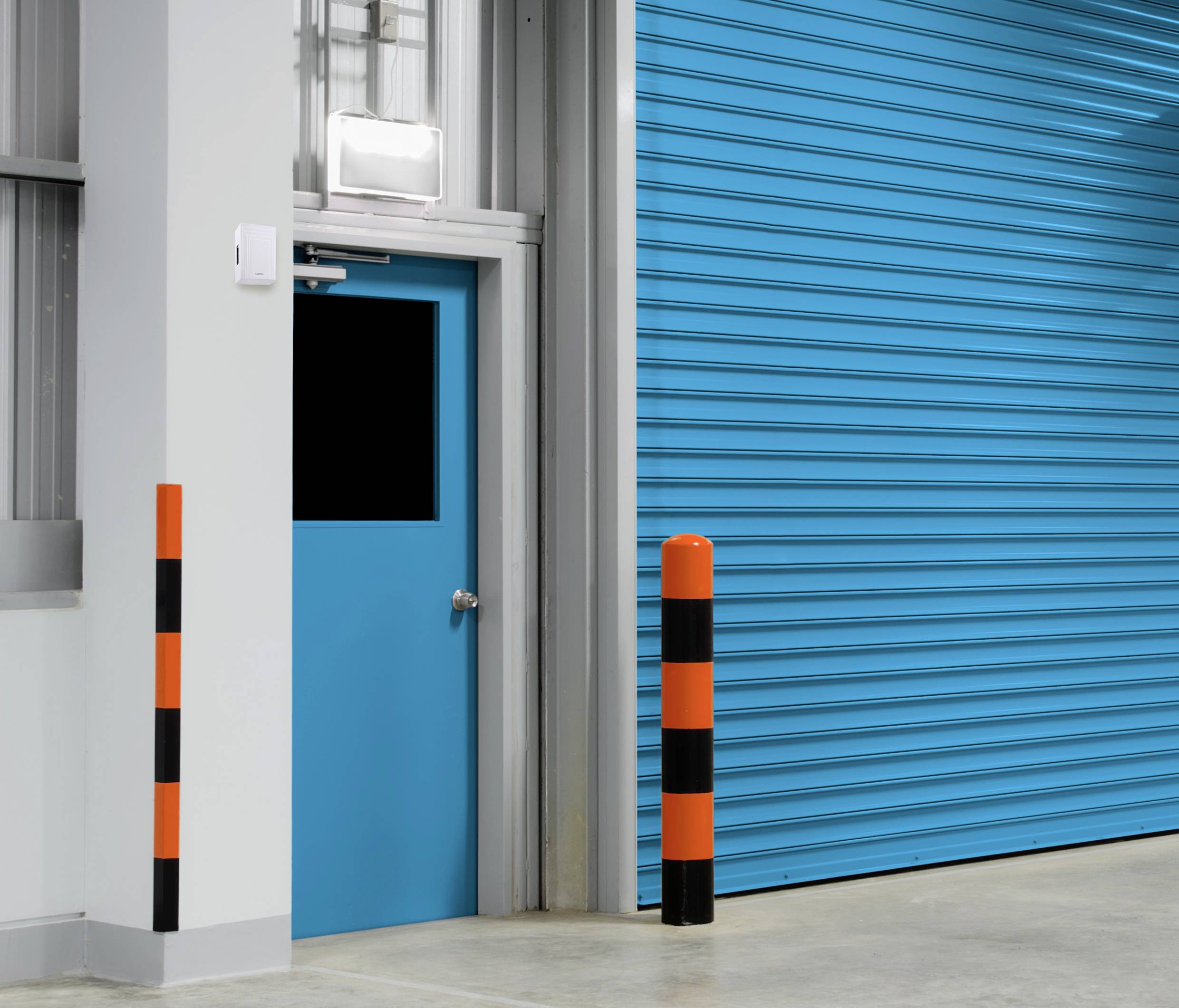 Modré dveře vedle velké, uzavřené, modré vratové mříže v průmyslovém objektu. V popředí stojí oranžovo-černé sloupky.