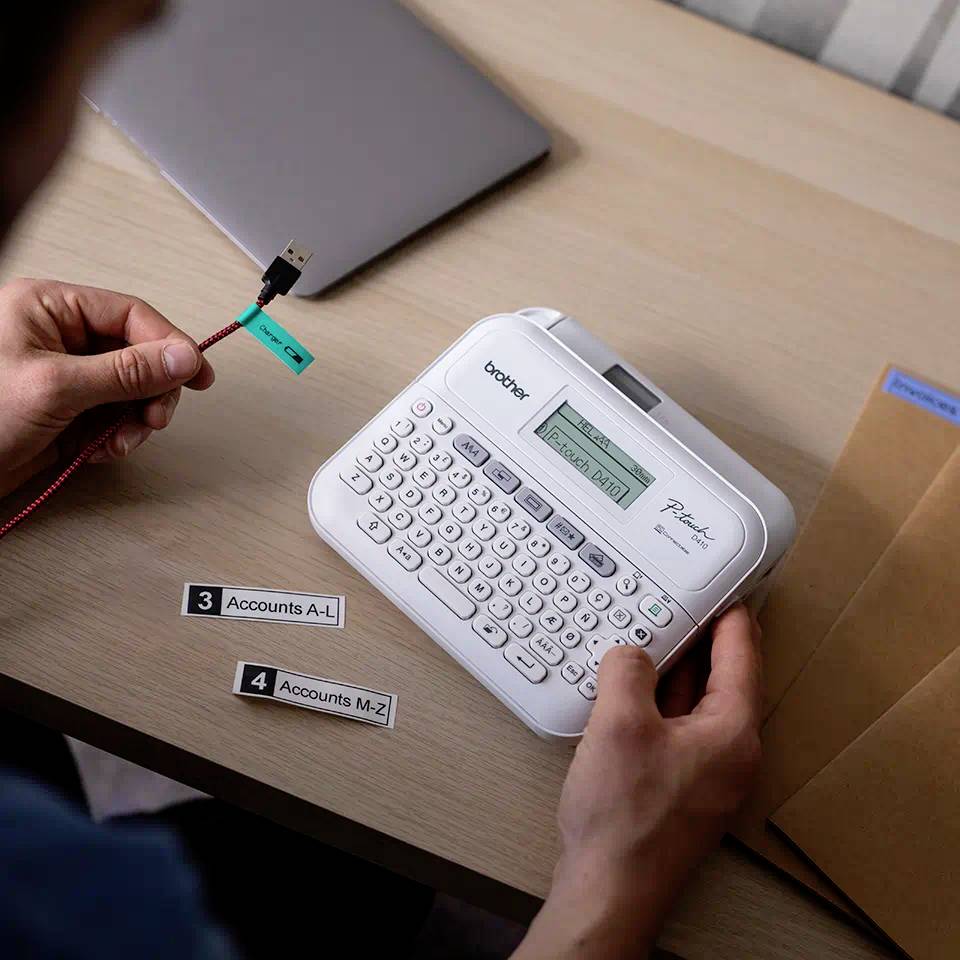 Jedna osoba používá zařízení na štítky, aby vytiskla etikety. Na stole leží notebook a obálky.