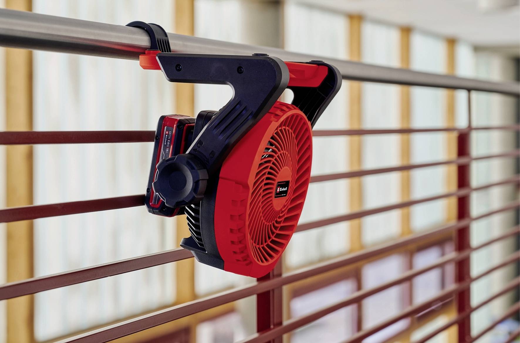 Červený přenosný ventilátor připevněný k zábradlí. V pozadí jsou vidět okna se světlými záclonami.