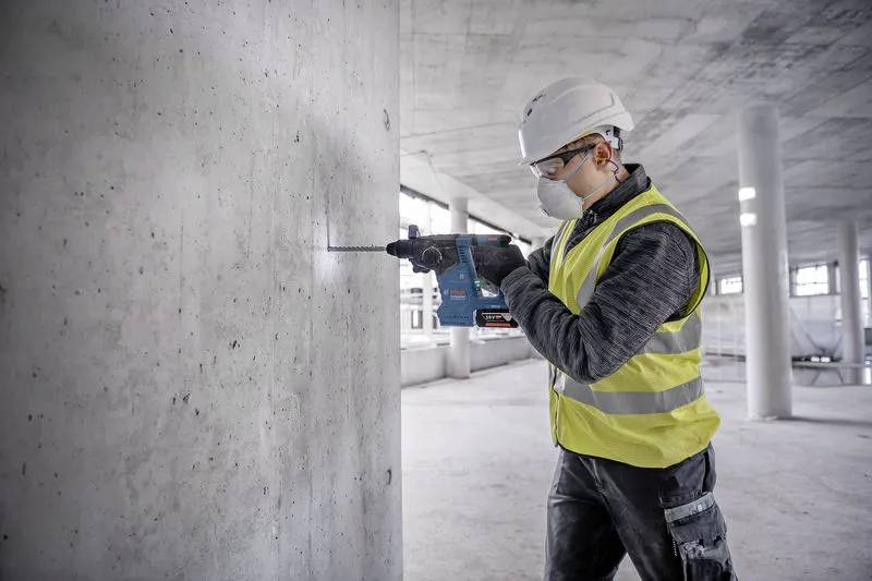Stavební dělník v přilbě a reflexní vestě používá elektrickou vrtačku na betonové stěně v nedokončeném interiéru budovy.