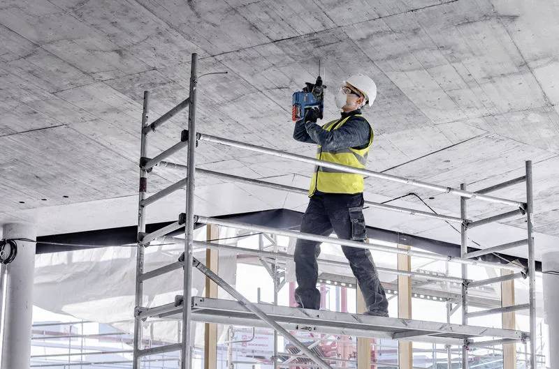 Dělník ve stavebním oblečení a bezpečnostní helmě stojí na lešení a vrtá do betonového stropu na staveništi.