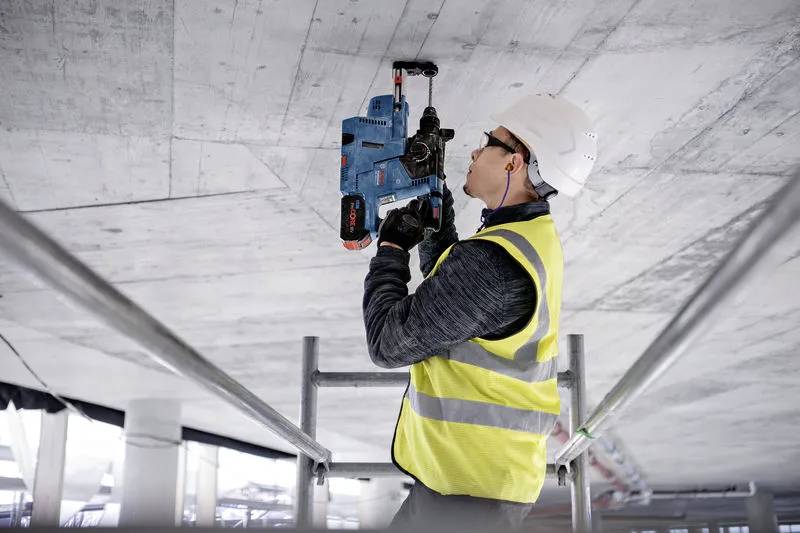 Dělník ve výstražné vestě a helmě používá elektrickou vrtačku, zatímco stojí na plošině a pracuje na betonové stropní konstrukci.
