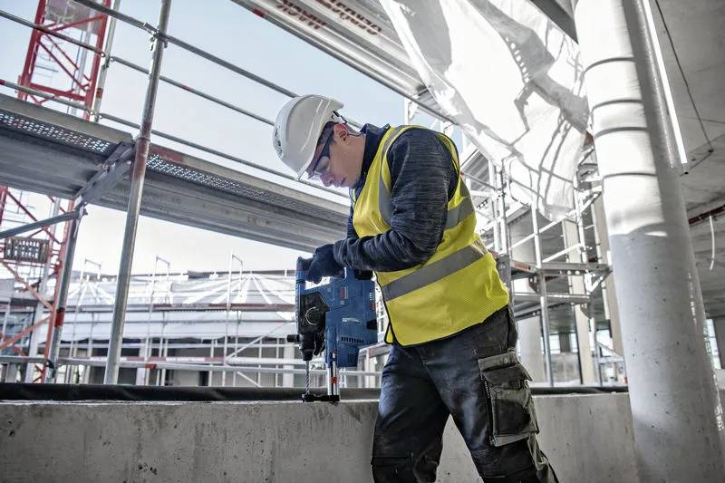 Dělník ve stavebnictví v žluté vestě a helmě používá elektrický nástroj na staveništi s lešením a konstrukční kostrou budovy.
