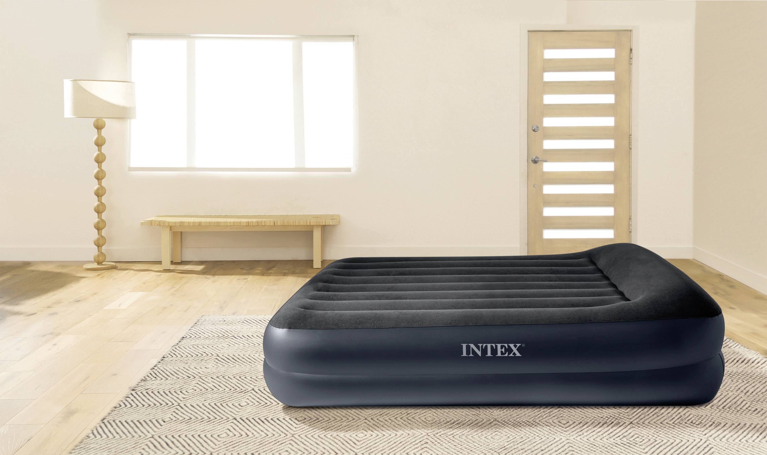 Nafukovací postel značky Intex stojí na koberci v světlém pokoji s lampou a oknem.