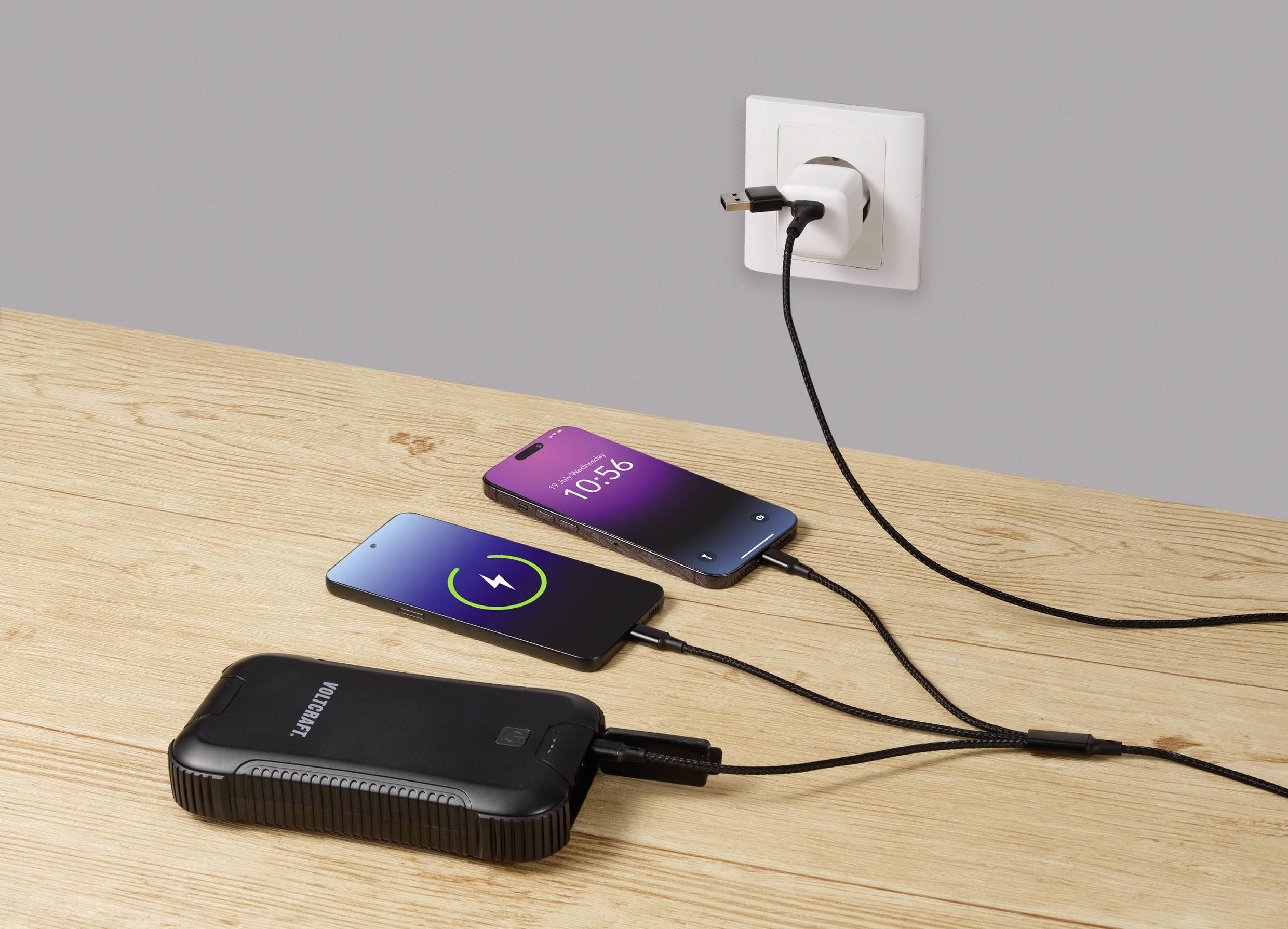 Dva smartphony leží na dřevěném stole a nabíjí se. Jeden zobrazuje zelený nabíjecí displej, druhý ukazuje čas 10:56. Jsou připojeny k elektrické zásuvce a powerbansce.