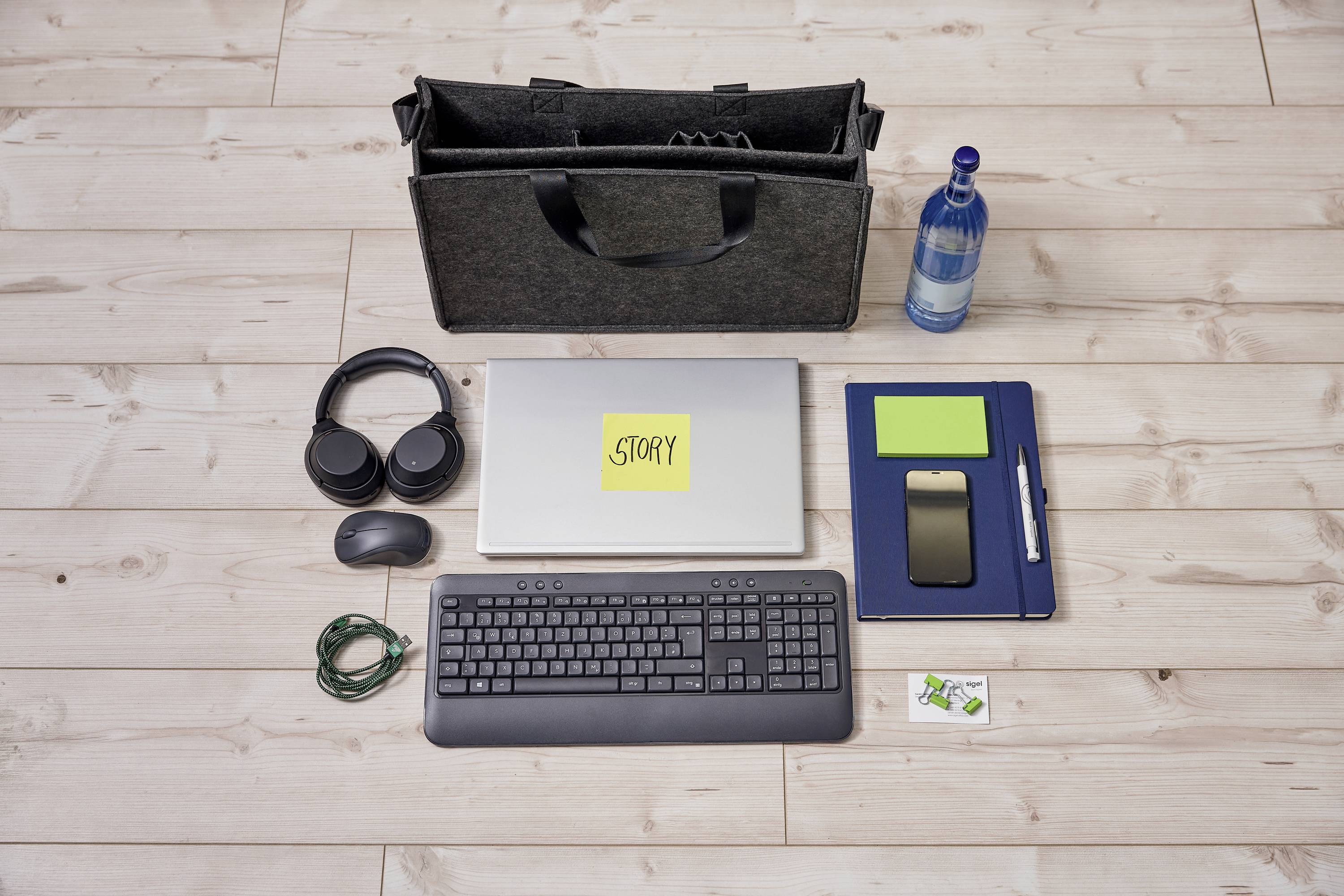 Laptop s žlutým lepicím lístkem 'Story' na víku, obklopený sluchátky, lahví, notes, mobilním telefonem, myší, kabely a klávesnicí.